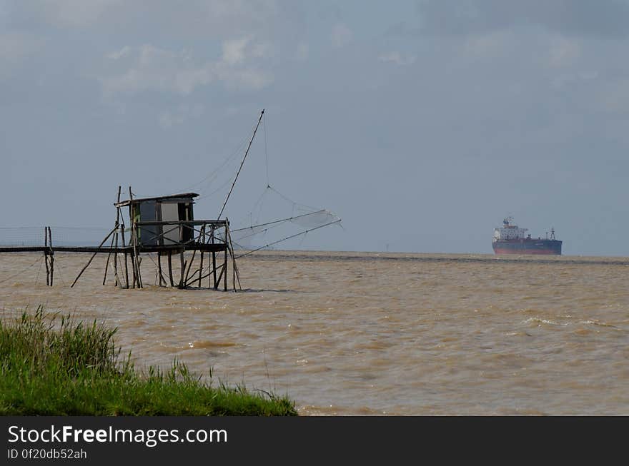 Wooden hut on dock in choppy waters with cargo ship on horizon.