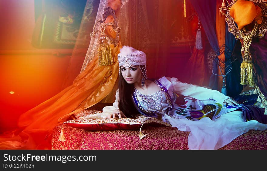 Beautiful young Arabic woman lying on opulent bedroom in traditional clothes with red tone. Beautiful young Arabic woman lying on opulent bedroom in traditional clothes with red tone.