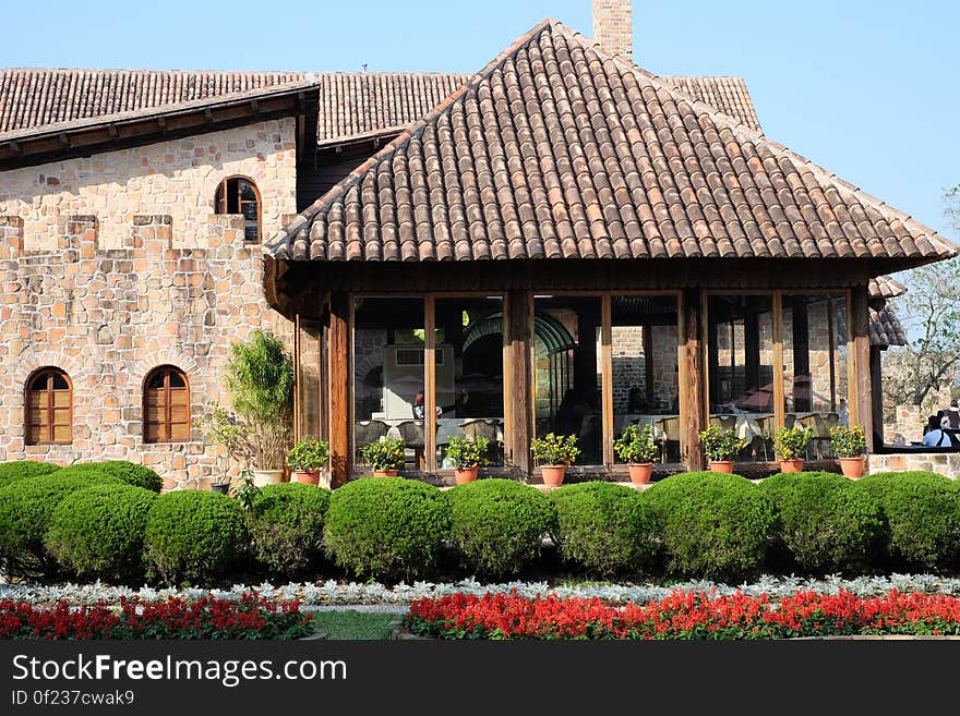 Exterior of a stone rustic building with conservatory, flowers and hedge in foreground.