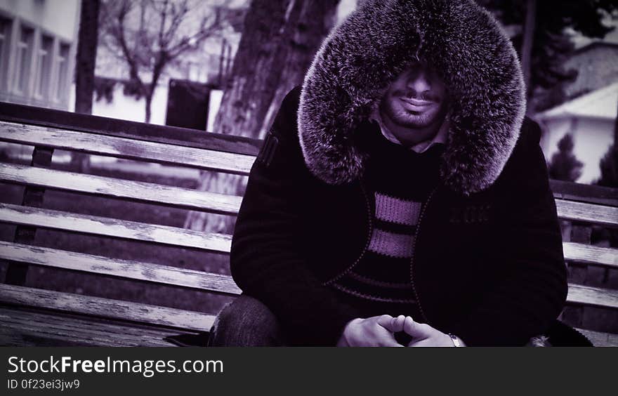 A man wearing a fur-lined jacket sitting on a bench. A man wearing a fur-lined jacket sitting on a bench.