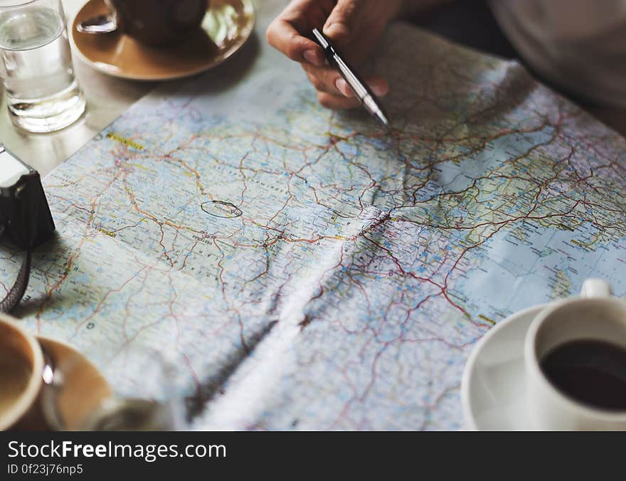 A roadmap on a table, person's hand holding a pen above, cups of coffee and a camera beside.