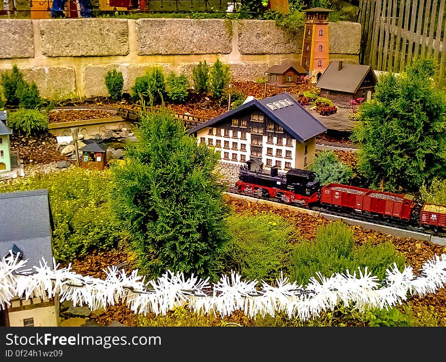 Miniature model railway with railway track and station in a garden with growing shrubs and trees and on the track a steam goods train with trucks filled with gray ballast.