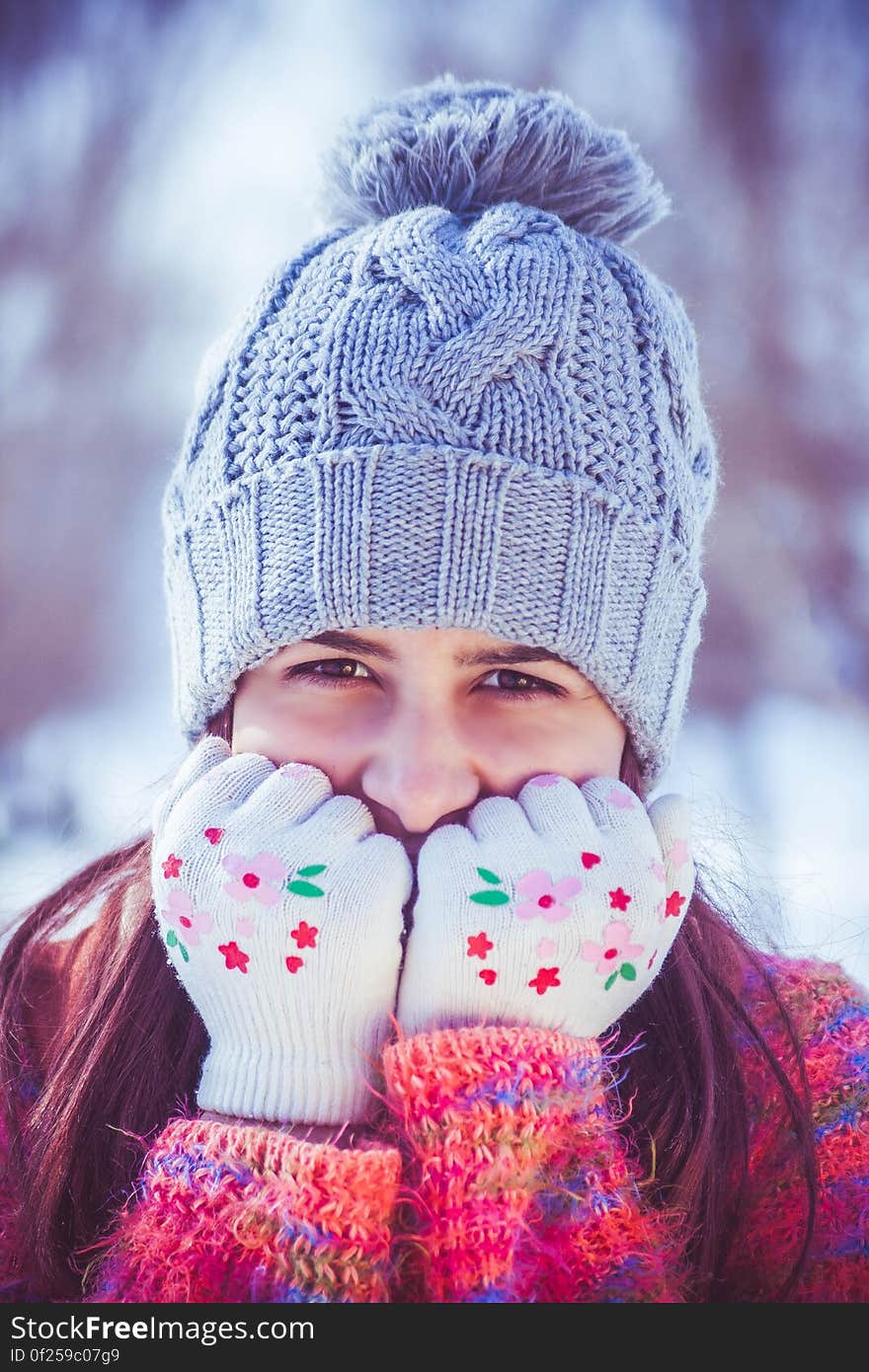 Portrait of pretty young woman in cold Winter weather wearing a gray colored knitted tea cozy hat with pom pom and red and purple jumper with gloved hands covering her mouth, mainly white background. Portrait of pretty young woman in cold Winter weather wearing a gray colored knitted tea cozy hat with pom pom and red and purple jumper with gloved hands covering her mouth, mainly white background.