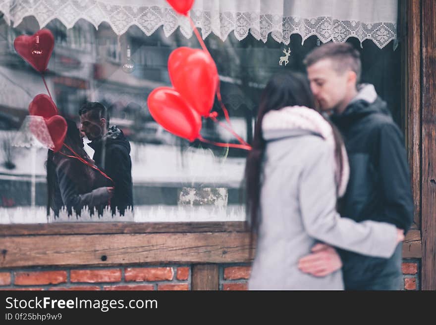 A couple holding heart shaped balloons kissing in front of a window. A couple holding heart shaped balloons kissing in front of a window.