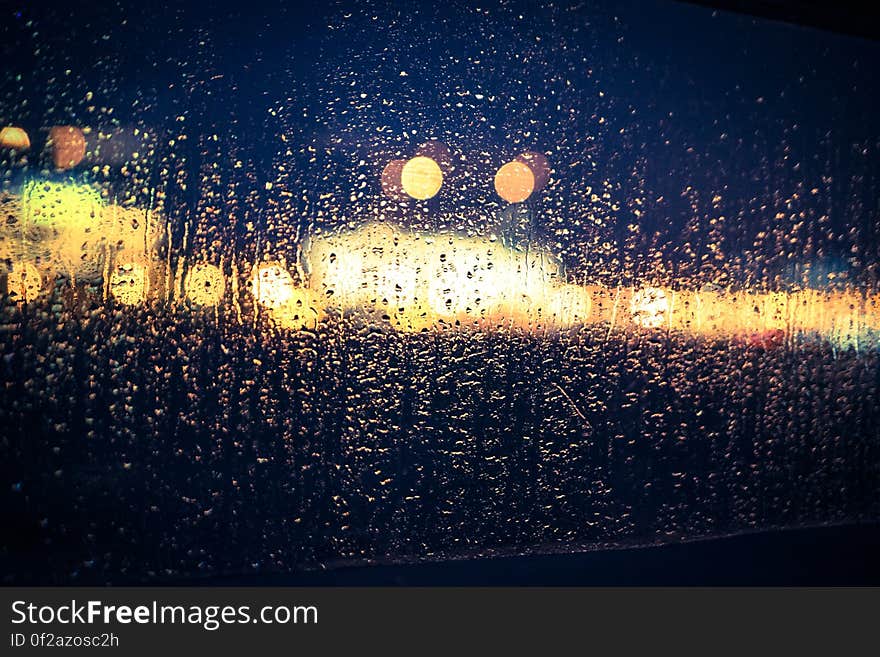 View through a window largely covered in rain drops to a road with traffic and a shopping mall showing up as a gold blur, black night background. View through a window largely covered in rain drops to a road with traffic and a shopping mall showing up as a gold blur, black night background.