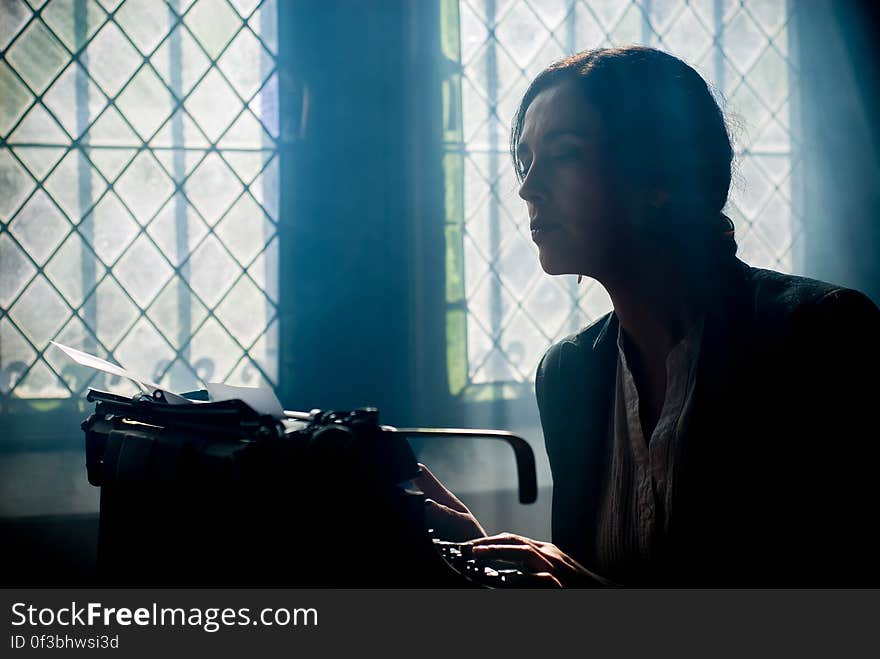 A woman typing on a typewriter by a window. A woman typing on a typewriter by a window.