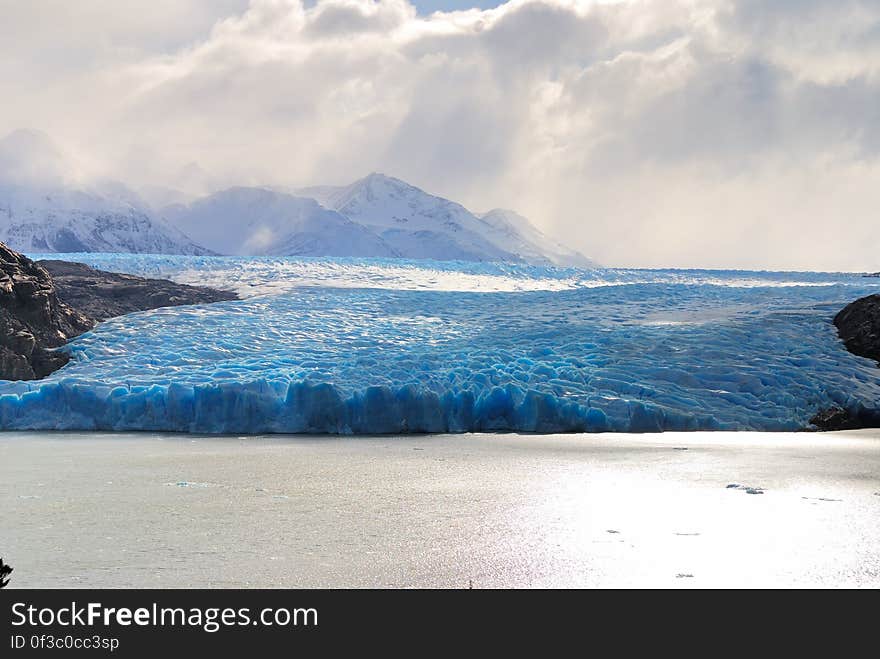 Edge of blue glacial ice along coastline with snow capped peaks against cloudy skies. Edge of blue glacial ice along coastline with snow capped peaks against cloudy skies.