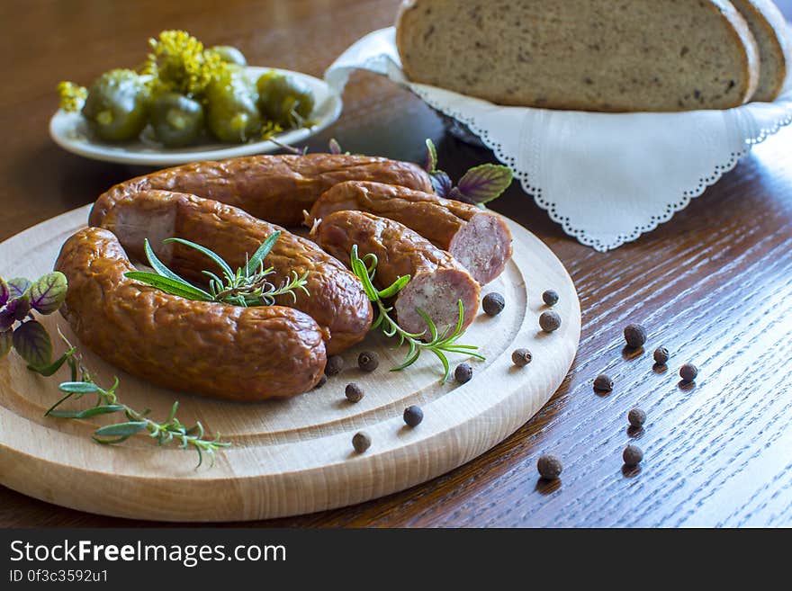 Close up of breakfast sausages with herbs and spices on wooden cutting board with basket of brown break ad bowl of pickles. Close up of breakfast sausages with herbs and spices on wooden cutting board with basket of brown break ad bowl of pickles.