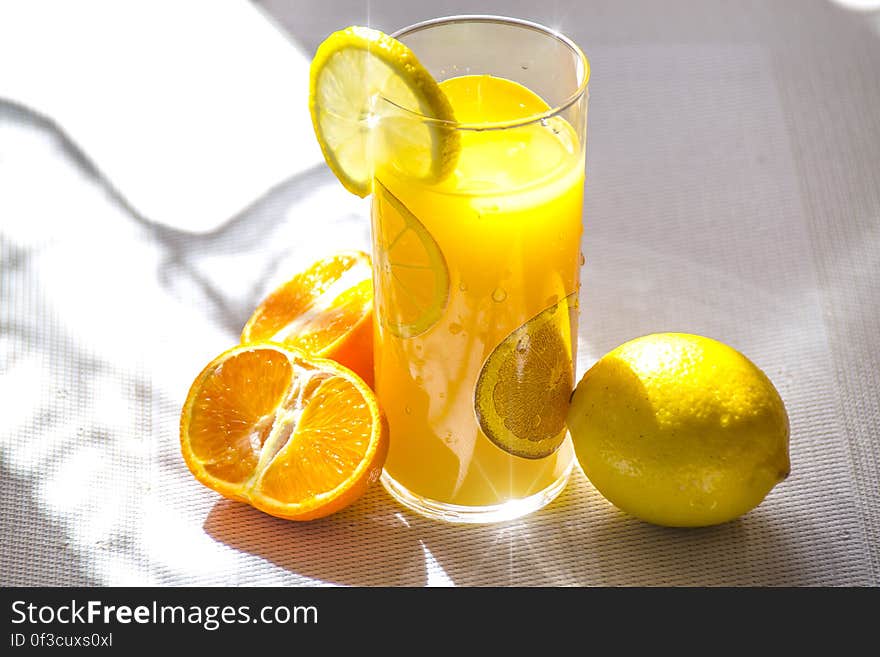 A glass with an yellow drink and lemon slice, lemon and orange next to it. A glass with an yellow drink and lemon slice, lemon and orange next to it.