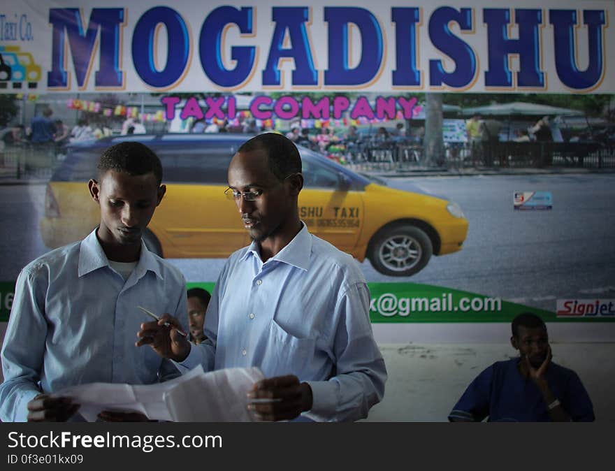 Suleiman Mohamed Daras &#x28;centre&#x29;, manager of the Mogadishu Taxi Company discusses fares and routes with a fellow worker at the company&#x27;s head office in the Somali capital Mogadishu, 01 September 2013. Operating since the end of May of this year in the city&#x27;s dusty and once again bustling streets and thoroughfares, the MTC&#x27;s distinctive yellow and purple cars offer customers taxi fares around Mogadishu at competitive rates and in the first 3 months of operations, the company has increased it&#x27;s fleet of vehicles from an initial 25 to over 100. The company, according to one of it&#x27;s drivers, also enables employment opportunities for Somalia&#x27;s youth following two decades of conflict in the Horn of Africa nation that shattered a generation. Now, thanks to the relative peace that has followed the departure of the Al-Qaeda-affiliated extremist group Al Shabaab from the city; an internationally recognised government for the first time in years and thousands of Diaspora Somalis returning home to invest in and rebuild their country, the MTC is one of many new companies establishing itself in the new Mogadishu and offering services that were hitherto impossible to provide. AU-UN IST PHOTO / STUART PRICE. Suleiman Mohamed Daras &#x28;centre&#x29;, manager of the Mogadishu Taxi Company discusses fares and routes with a fellow worker at the company&#x27;s head office in the Somali capital Mogadishu, 01 September 2013. Operating since the end of May of this year in the city&#x27;s dusty and once again bustling streets and thoroughfares, the MTC&#x27;s distinctive yellow and purple cars offer customers taxi fares around Mogadishu at competitive rates and in the first 3 months of operations, the company has increased it&#x27;s fleet of vehicles from an initial 25 to over 100. The company, according to one of it&#x27;s drivers, also enables employment opportunities for Somalia&#x27;s youth following two decades of conflict in the Horn of Africa nation that shattered a generation. Now, thanks to the relative peace that has followed the departure of the Al-Qaeda-affiliated extremist group Al Shabaab from the city; an internationally recognised government for the first time in years and thousands of Diaspora Somalis returning home to invest in and rebuild their country, the MTC is one of many new companies establishing itself in the new Mogadishu and offering services that were hitherto impossible to provide. AU-UN IST PHOTO / STUART PRICE.