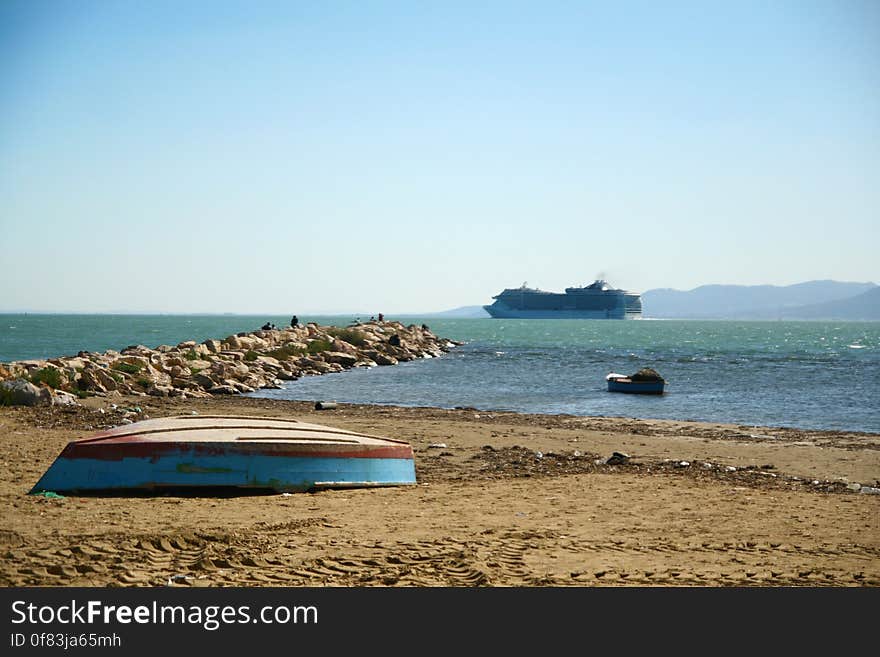 Blue, wooden boat, upside down, lying on an empty beach, by the sea, far away in the sea swims a large cruise ship