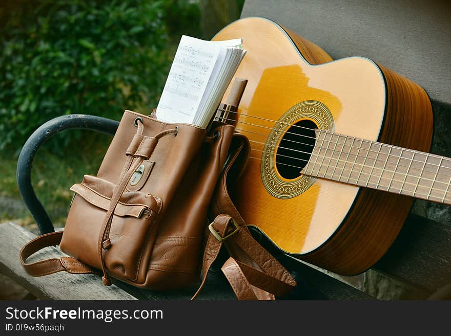 Brown Acoustic Guitar Beside Brown Leather Bucket Backpack on Brown Wooden Bench