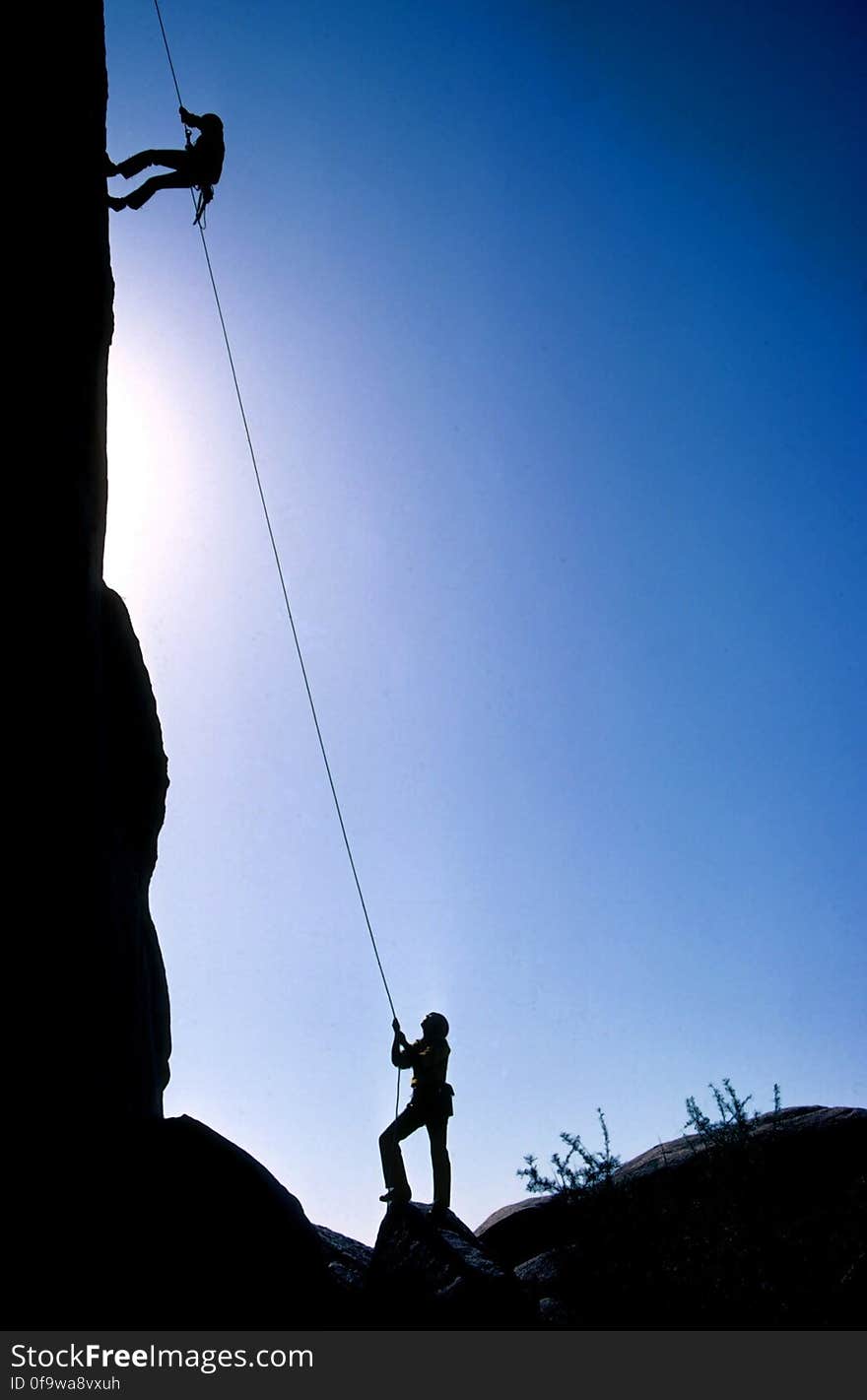 Silhouette of mountain climbers scaling cliff face against blue skies. Silhouette of mountain climbers scaling cliff face against blue skies.