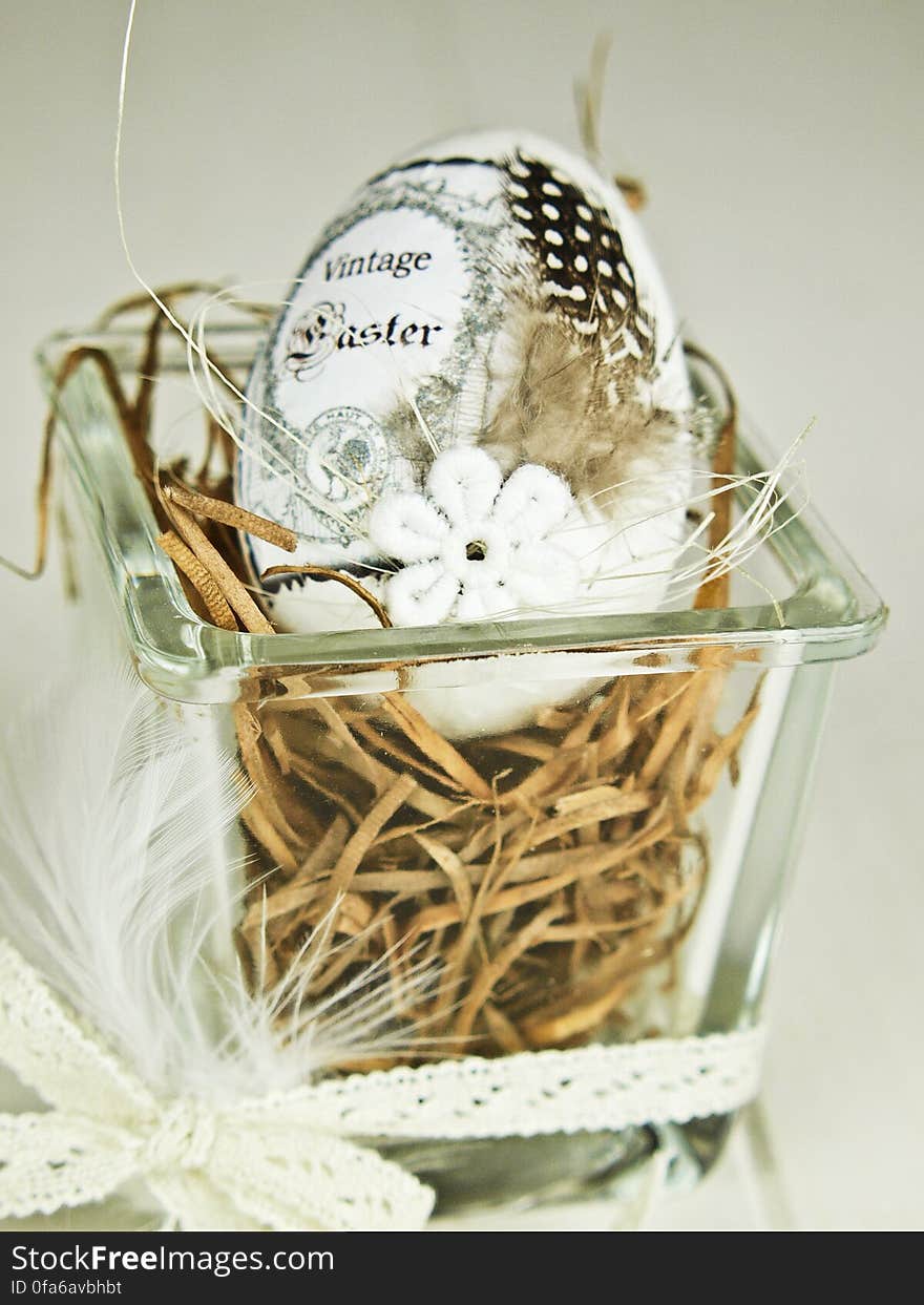 A close up of a traditional Easter egg on straw in a transparent square bowl decorated with white ribbon.