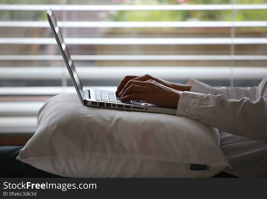 Person Wearing White Dress Shirt Using Silver Laptop on Top of White Throw Pillow