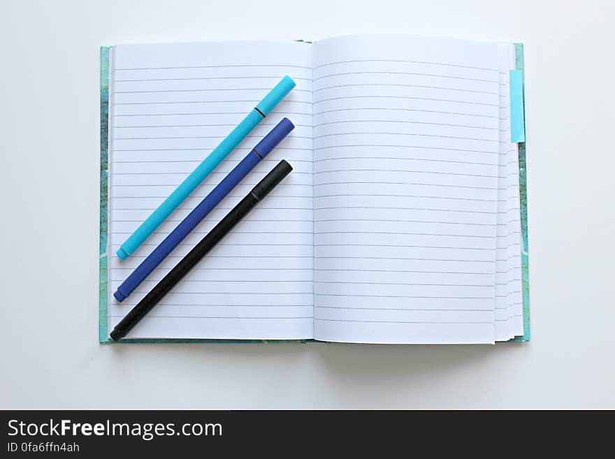 Open lined notebook open with three colored pens lying diagonally across one page, white background. Open lined notebook open with three colored pens lying diagonally across one page, white background.