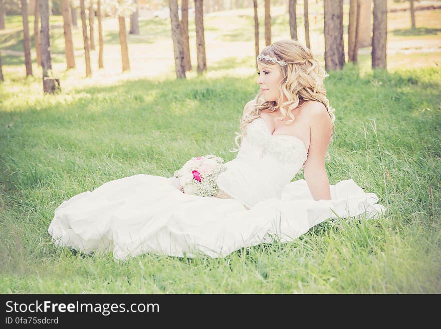 Young blond haired bride sitting in green field with bouquet on sunny day. Young blond haired bride sitting in green field with bouquet on sunny day.