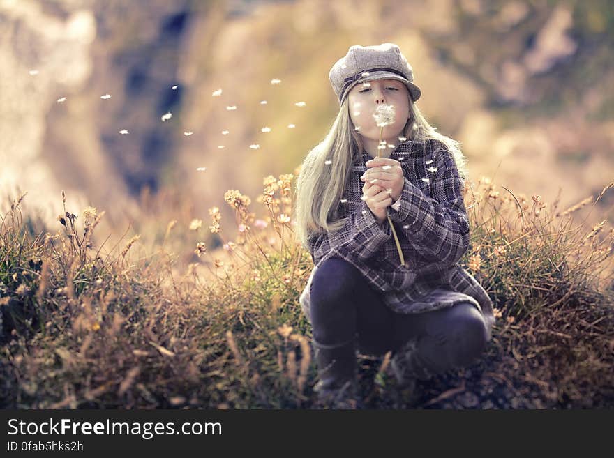 Girl in plaid jacket an hat kneeling in field blowing dandelions. Girl in plaid jacket an hat kneeling in field blowing dandelions.