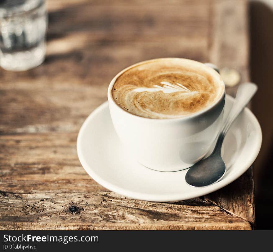 Coffee latte in a white cup and saucer placed near the edge of a rough wooden table, glass of water nearby. Coffee latte in a white cup and saucer placed near the edge of a rough wooden table, glass of water nearby.