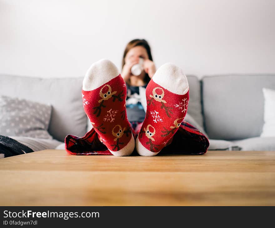 Girl lying on a couch drinking from a white mug and wearing slippers with Christmas style decorations. Selective focus in on the soles of the slippers. Girl lying on a couch drinking from a white mug and wearing slippers with Christmas style decorations. Selective focus in on the soles of the slippers.