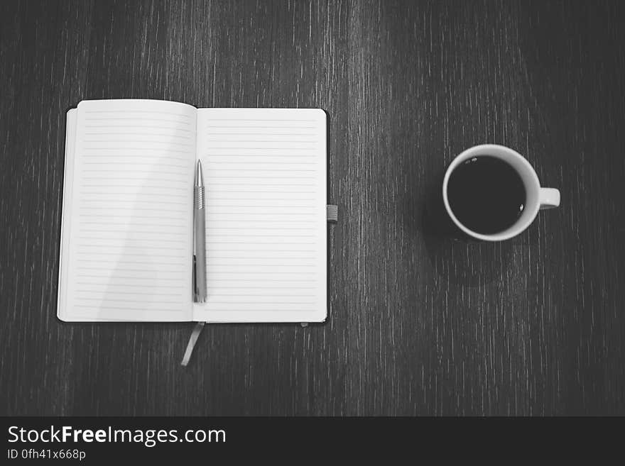 Black and white photo of an open blank notebook with a pen and a cup of coffee next to it on a wooden table. Black and white photo of an open blank notebook with a pen and a cup of coffee next to it on a wooden table.