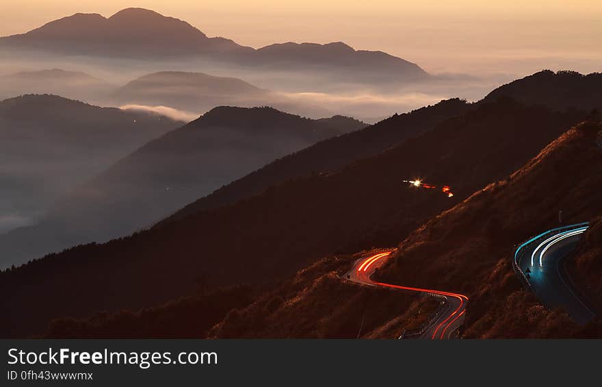 Cars zigzagging along a dark mountain road with headlights and rear lights highlighting the road at dawn producing continuous red and yellow tracks. Cars zigzagging along a dark mountain road with headlights and rear lights highlighting the road at dawn producing continuous red and yellow tracks.