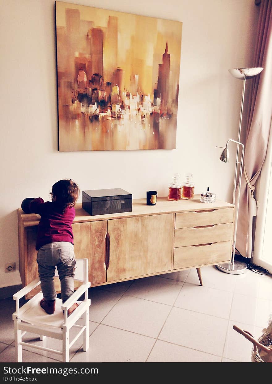 Child climbing on chair in living room by cabinet with objects on top.