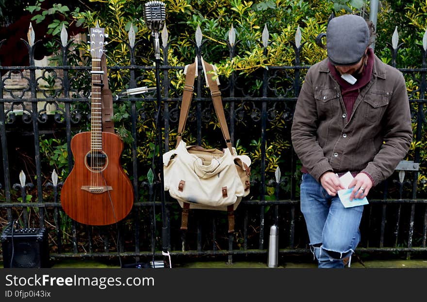 Man Leaning Against Black Steel Fence Beside White and Brown Sling Bag and Brown Acoustic Guitar