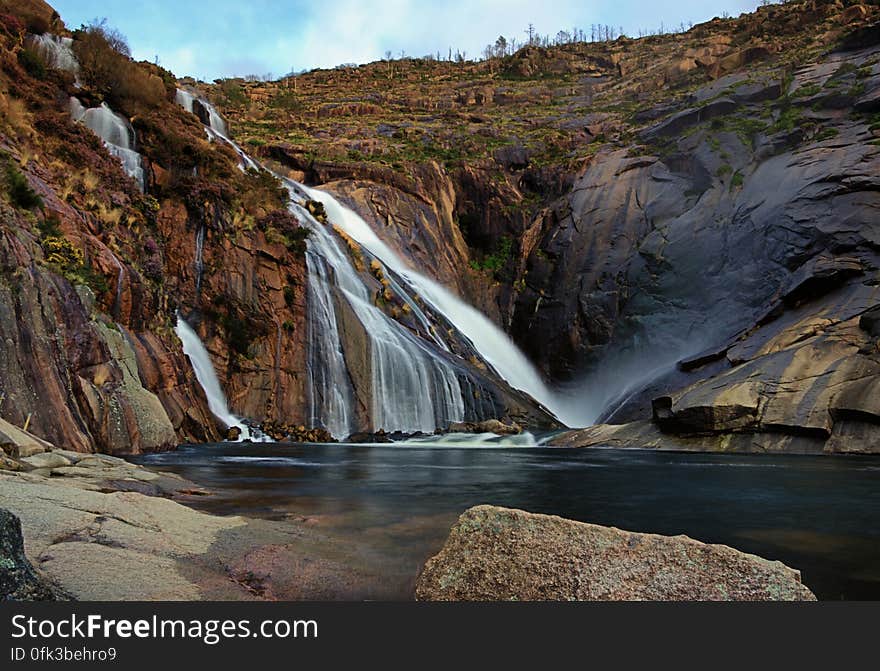 Waterfall over rocky hillside into clear pool in mountain landscape.