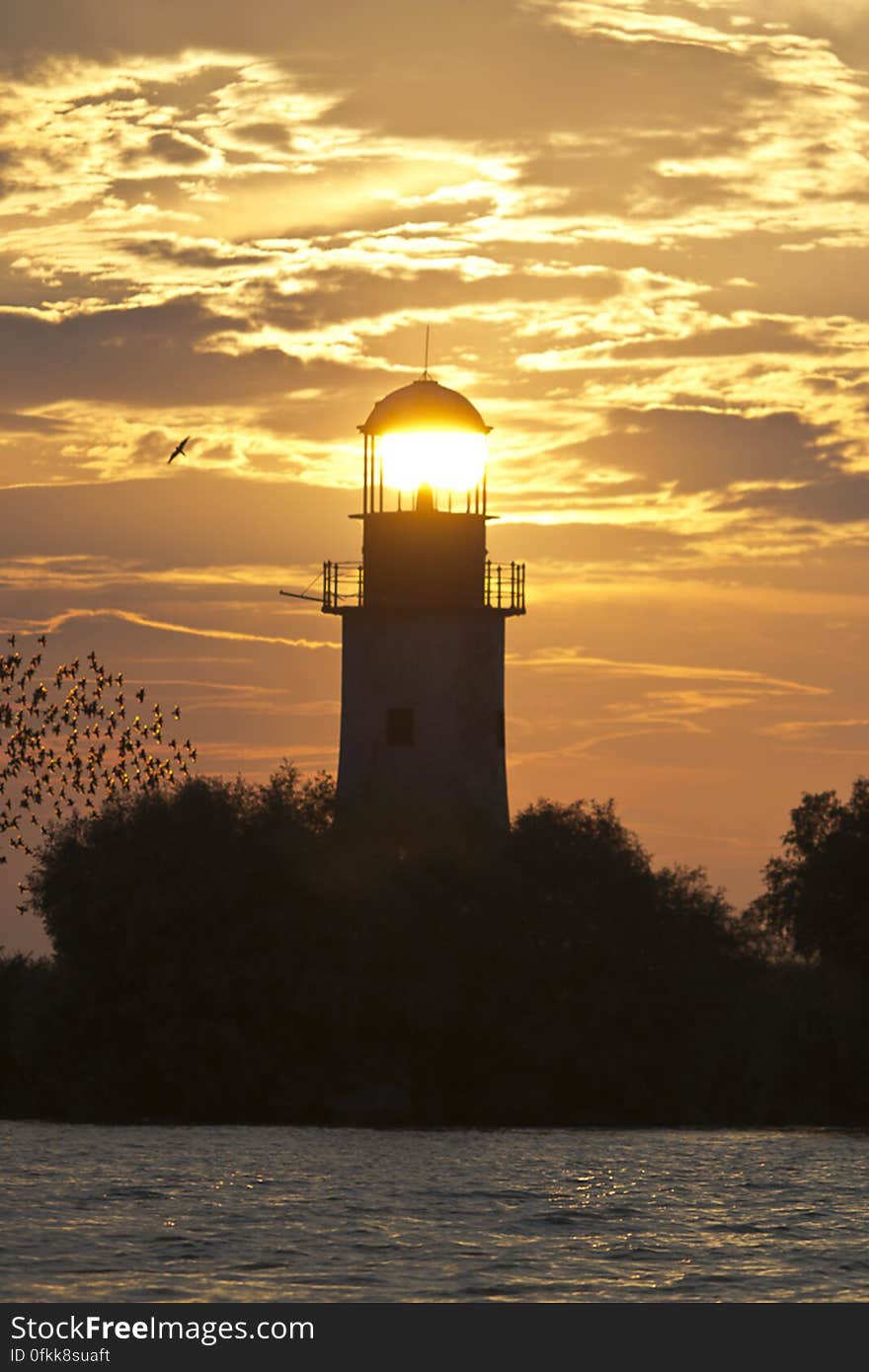 Setting sun seems to lit an old lighthouse while a flock of starlings fly against the orange colored sky. Setting sun seems to lit an old lighthouse while a flock of starlings fly against the orange colored sky.