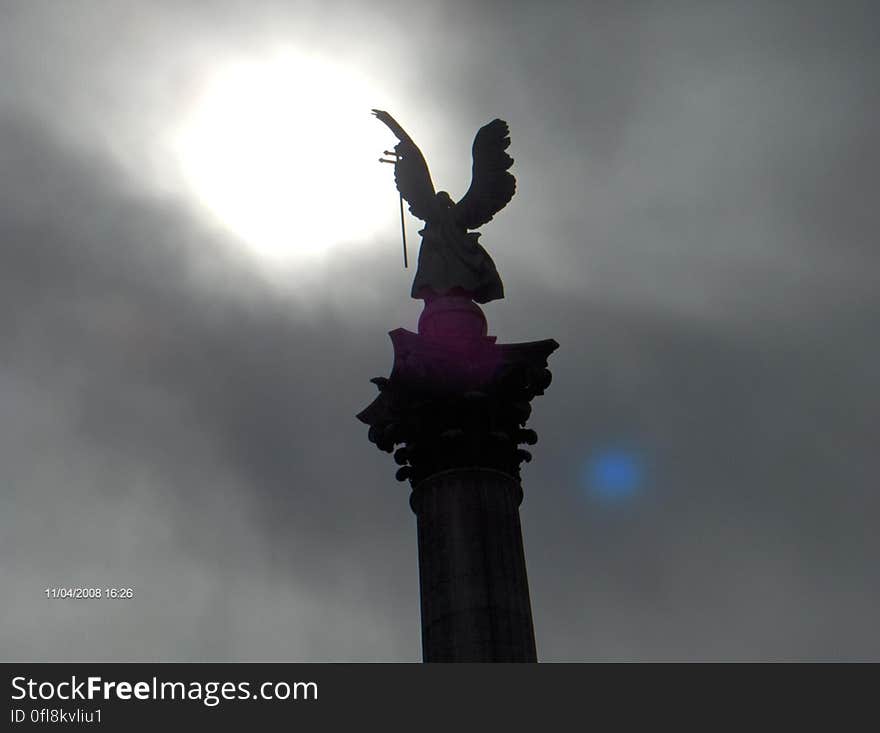 A silhouetted angel statue and the sun breaking through clouds in the background.