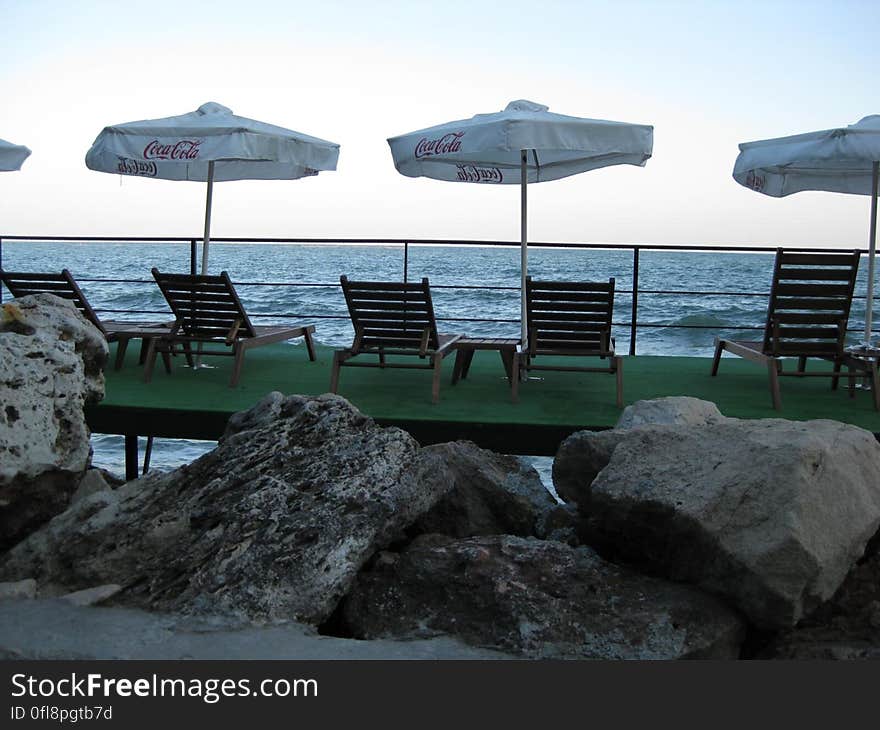 Loungers for holiday makers at the seaside in front of large rocks with shade provided by white parasols advertising Coca Cola, pale blue sky. Loungers for holiday makers at the seaside in front of large rocks with shade provided by white parasols advertising Coca Cola, pale blue sky.