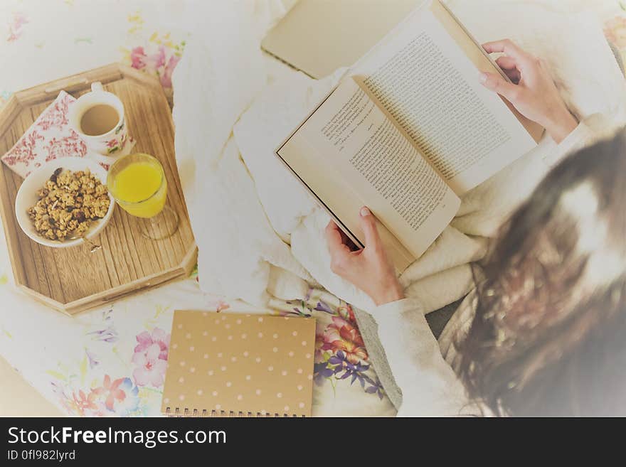 A young woman reading a book in bed, juice, coffee and crisps next to her on a tray. A young woman reading a book in bed, juice, coffee and crisps next to her on a tray.