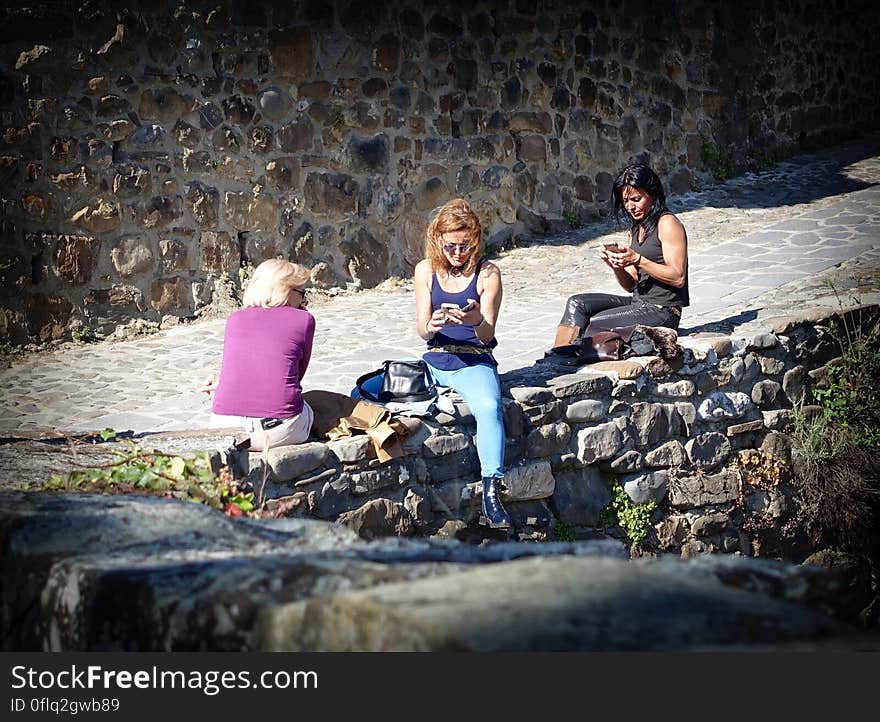 A group of people sitting on a stone wall in the sun looking at their phones. A group of people sitting on a stone wall in the sun looking at their phones.