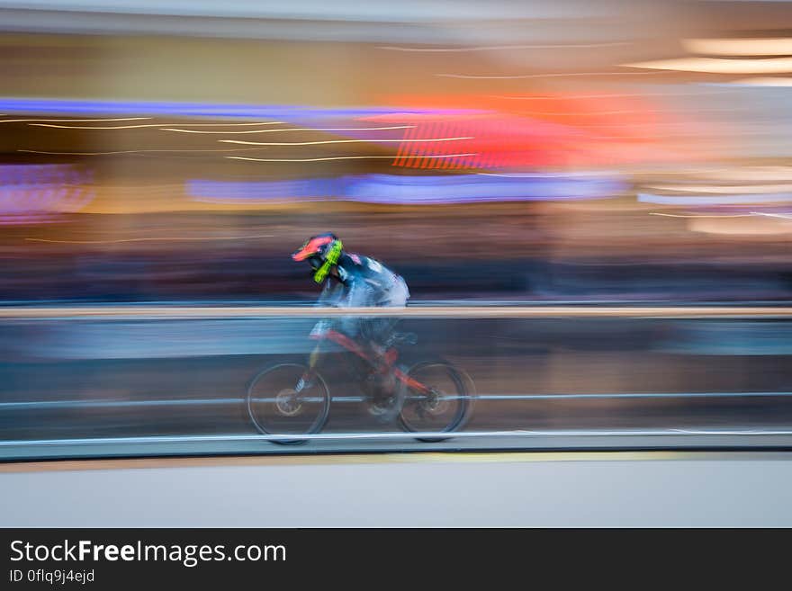 Biker on red bicycle wearing red helmet and blue jersey streaks past colorful buildings and beyond them a high mountain. Biker on red bicycle wearing red helmet and blue jersey streaks past colorful buildings and beyond them a high mountain.