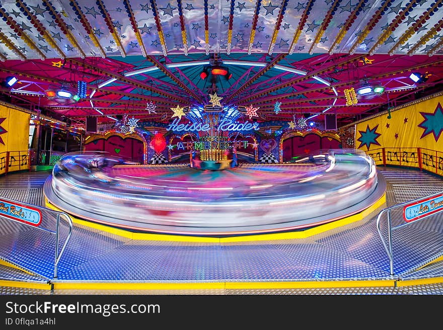 Roundabout marked above it by "Heisse Raider" in rapid motion creating blur, at a colorful funfair. Roundabout marked above it by "Heisse Raider" in rapid motion creating blur, at a colorful funfair.