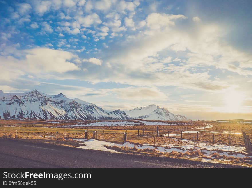 Snow covered mountain range next to country field with empty road on sunny day. Snow covered mountain range next to country field with empty road on sunny day.