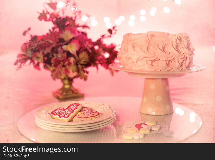 Cake on serving dish with plate of heart shaped cookies next to floral arrangement with pink background. Cake on serving dish with plate of heart shaped cookies next to floral arrangement with pink background.