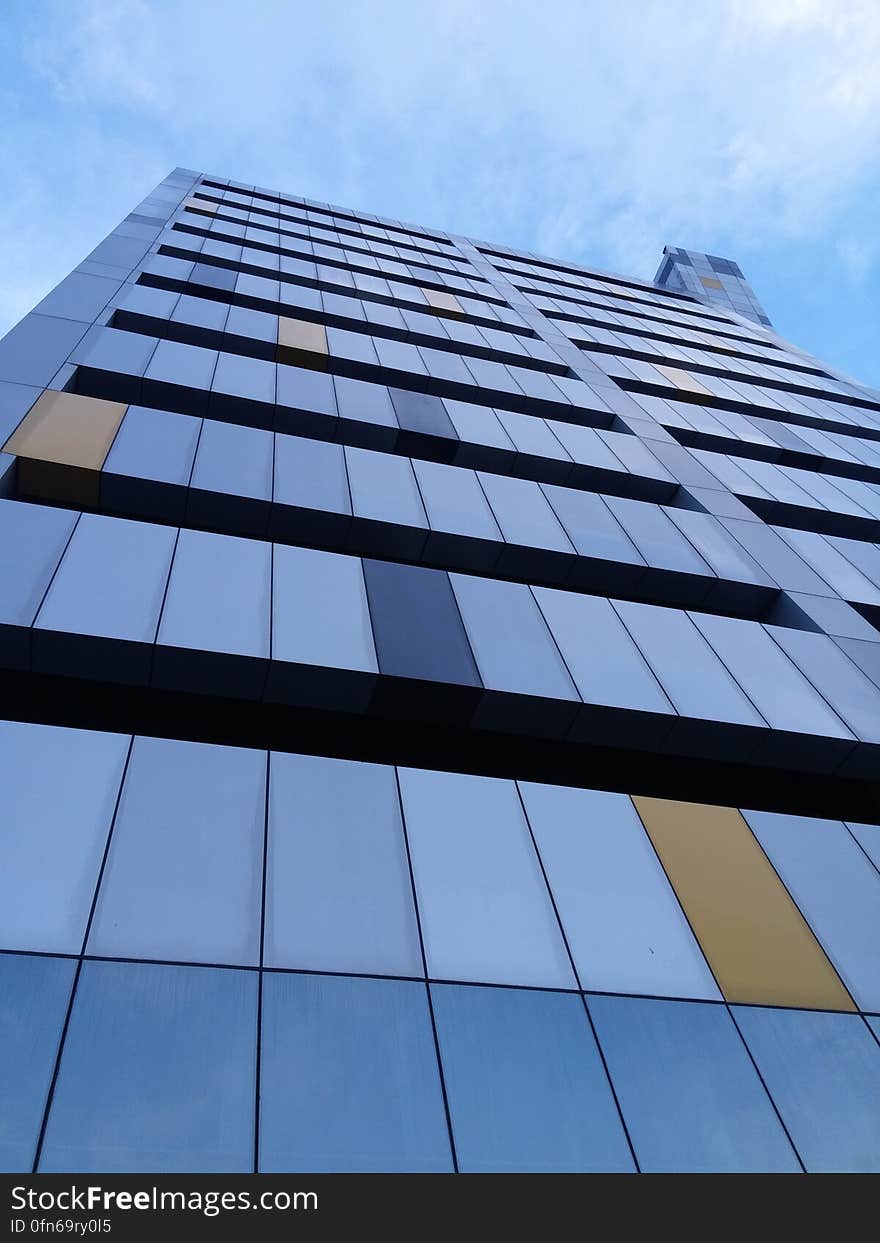 Slabs in gray, shades of blue and brown used on one face of a high rise building with dark opaque glass forming a band on each floor, pale blue sky. Slabs in gray, shades of blue and brown used on one face of a high rise building with dark opaque glass forming a band on each floor, pale blue sky.