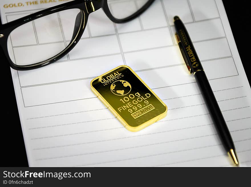 Still life of gold bar ingot with pen and black eyeglasses on blank sheet of paper. Still life of gold bar ingot with pen and black eyeglasses on blank sheet of paper.