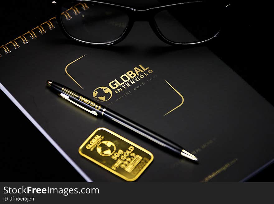 Still life with notebook stamped with Global Intergold logo and text with pen, eyeglasses and gold ingot. Still life with notebook stamped with Global Intergold logo and text with pen, eyeglasses and gold ingot.