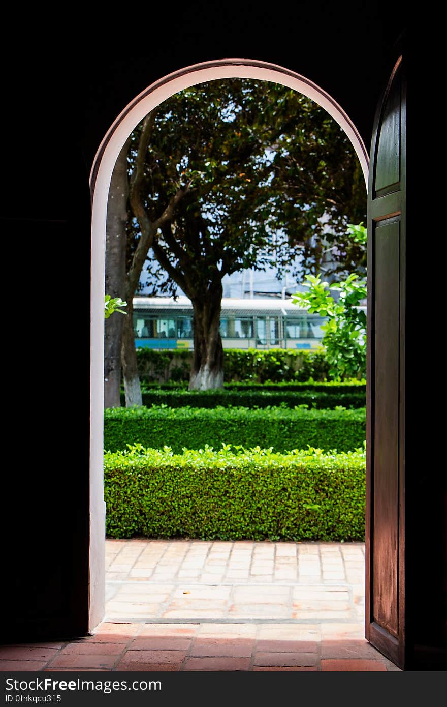 Open wooden door showing green hedges and trees next to urban street with bus. Open wooden door showing green hedges and trees next to urban street with bus.