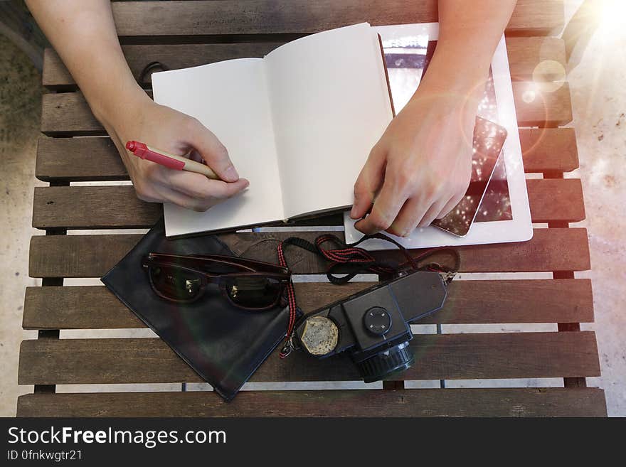 Two disembodied hands, one holding pen writing in a notebook placed on a slatted chair or table surrounded by camera, sun glasses, mobile phone and black case. Two disembodied hands, one holding pen writing in a notebook placed on a slatted chair or table surrounded by camera, sun glasses, mobile phone and black case.