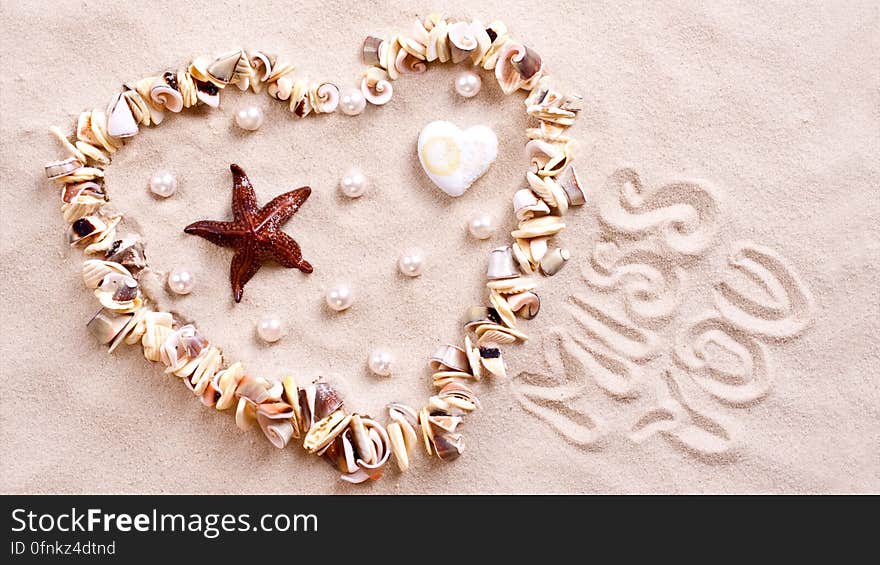 Seashell heart in sand with miss you spelled out.