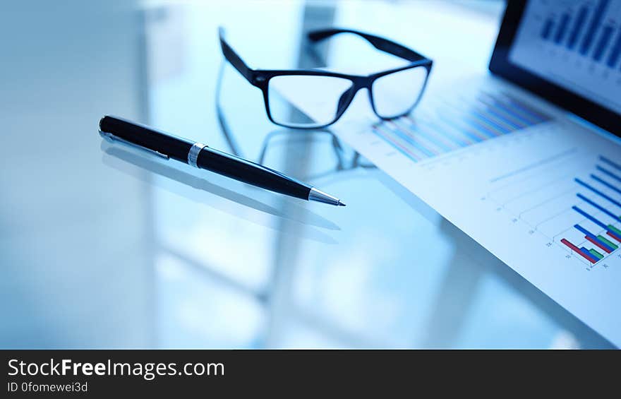 Glasses and pen on blue toned table next to data on business documents and computer screen. Glasses and pen on blue toned table next to data on business documents and computer screen.