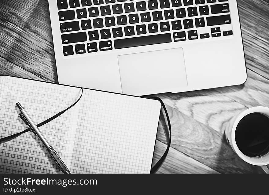 A black and white photo of laptop, notebook, and cup of coffee on a table. A black and white photo of laptop, notebook, and cup of coffee on a table.