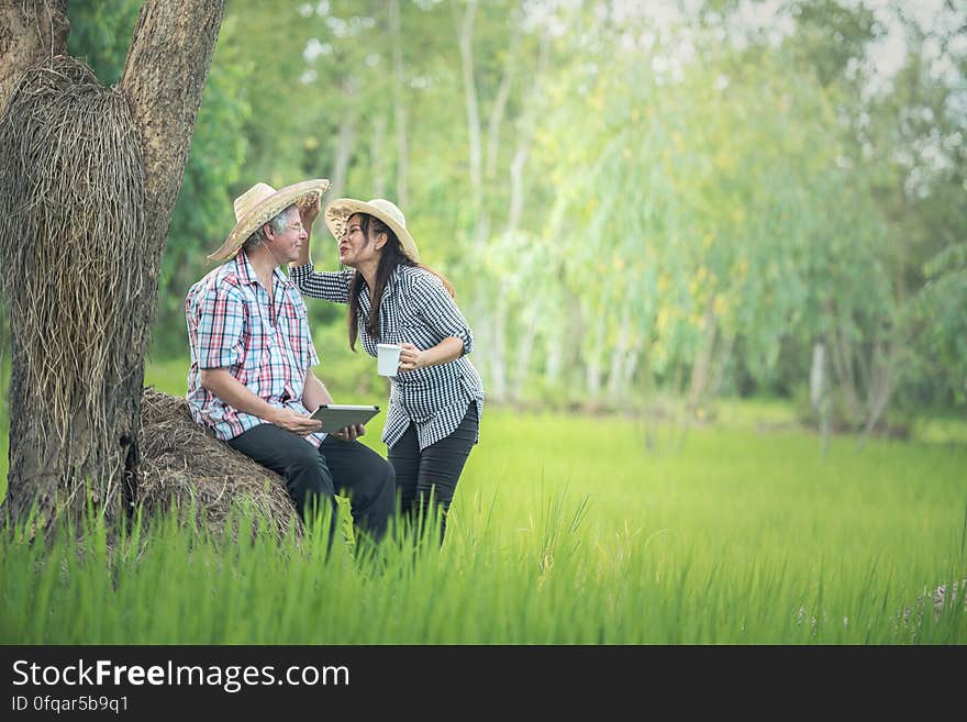 A man sitting on a tree trunk with a tablet in hand and an Asian woman holding a cup kissing him. A man sitting on a tree trunk with a tablet in hand and an Asian woman holding a cup kissing him.