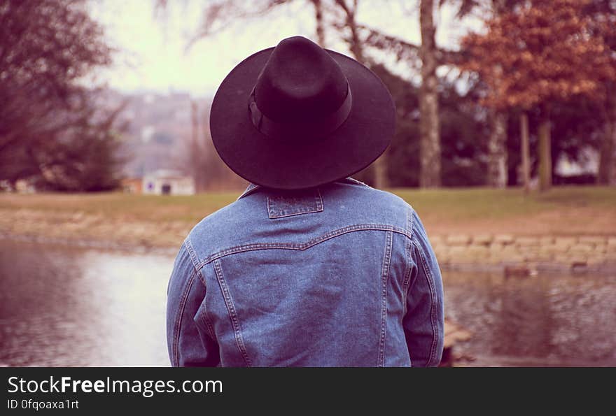 A person wearing a denim jacket and a fedora by a pond in a park. A person wearing a denim jacket and a fedora by a pond in a park.