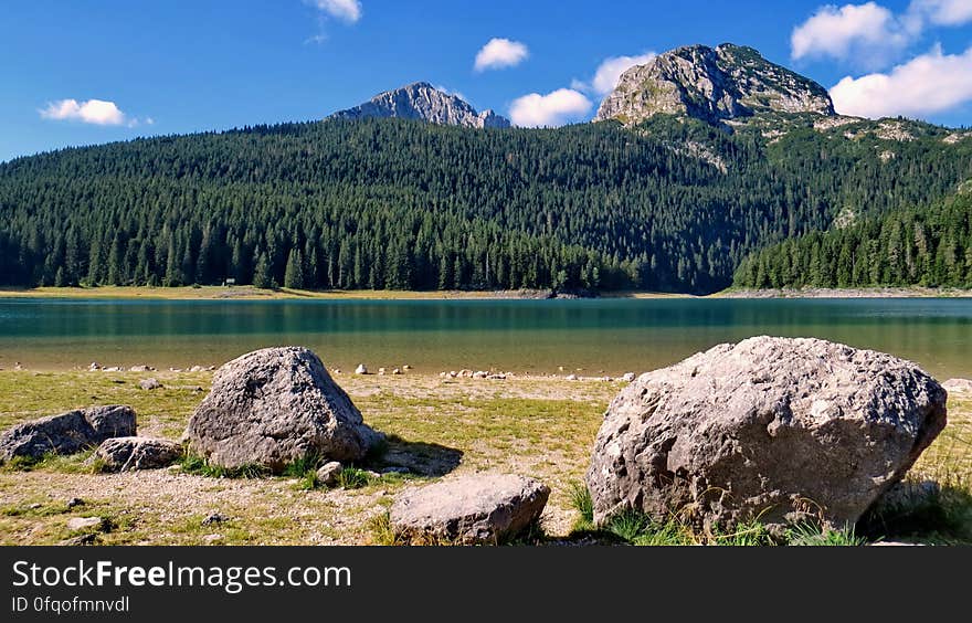 Large rocks on the shore of a lake on the far side of which is a dense fir forest, mountain peaks, blue sky and cloud background. Large rocks on the shore of a lake on the far side of which is a dense fir forest, mountain peaks, blue sky and cloud background.