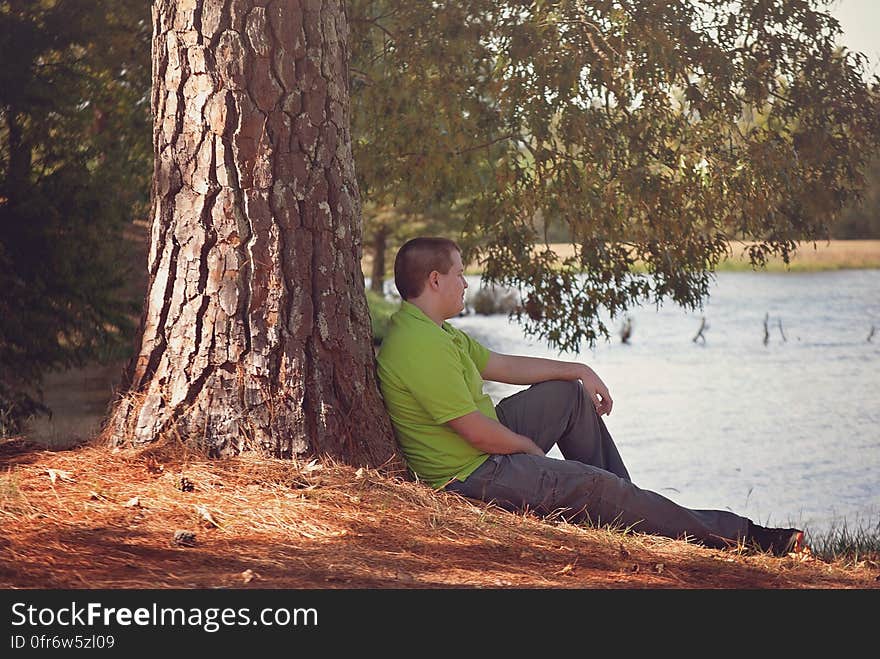 A man with his back against a tree looking at water. A man with his back against a tree looking at water.